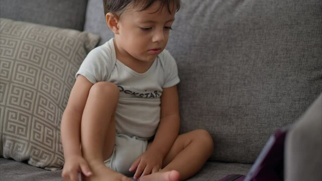 Nice latin toddler in a light blue baby romper sitting on a grey couch watching cartoons on a purple tablet