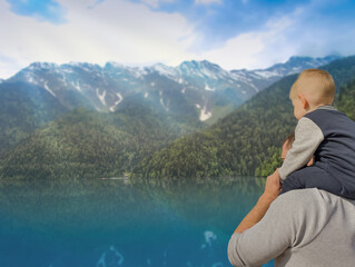 Fototapeta na wymiar A man with a small child stand on the shore of a mountain lake and look at the mountains. Dad and son. The child is sitting on Dad's shoulders. Family. View from the back. Summer, autumn day.