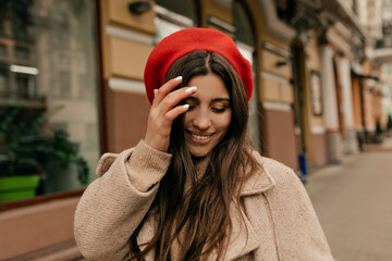 Close up portrait of lovely cute woman touching her hair, looking down and smiling. She wears red beret, beige coat 