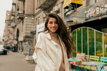 Happy excited lovely girl with wonderful smile wearing white trench and sweater posing at camera while walking on the street 