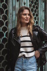 Young street fashion blond woman posing against vintage wooden door. Trendy style, black leather jacket, blue jeans, trendy autumn fashion collection. Three quarters portrait crop.