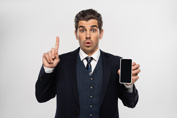 surprised businessman in suit holding smartphone with blank screen and pointing up isolated on grey