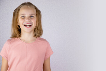 Portrait of a cute girl on a light background, a little blonde looks at the camera and smiles broadly. The concept of joy, happiness and good mood. Copyright.