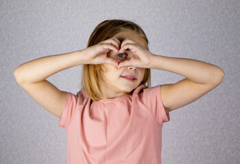 Portrait of a cute little blonde girl on a light background. She shows heart gesture with her hands