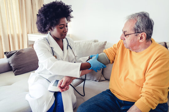 Healthcare Worker Taking Blood Pressure Of Senior Man At Home. Woman Doctor Cardiologist Or Physician Checking High Blood Pressure Examining Male Patient Using Tonometer During Home Medical Care Visit