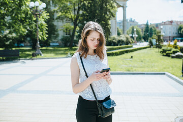 Happy beautiful young Caucasian girl with smart phone outdoors on sunny summer day texting and smiling