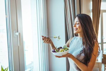 Healthy young woman dieting and eating a salad at home. Attractive woman with fresh green salad at home. A beautiful young woman is eating a salad in her living room, sitting on a sofa