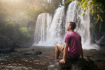 Man resting on stone in front of high waterfall in mountains in tropical landscape in Camobodia.