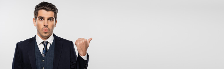 surprised businessman in suit pointing with thumb away isolated on grey, banner