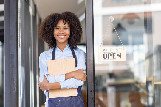 Happy waitress standing at restaurant entrance. Portrait of african american business woman attend new customers in her coffee shop. Happy woman owner showing open sign in her small business shop.