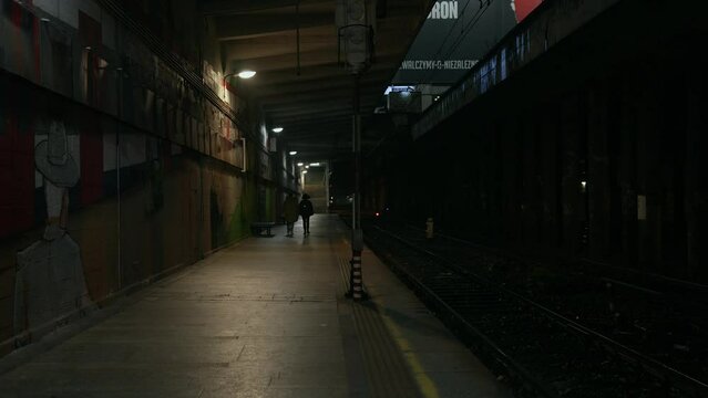Abandoned railway / subway platform. Ghetto. Rainy autumn foggy weather in the night city. Gotham city mood. Cinematic style. 
Two people are walking along the platform of a railway station. 