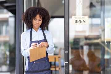 Happy waitress standing at restaurant entrance. Portrait of african american business woman attend new customers in her coffee shop. Happy woman owner showing open sign in her small business shop.