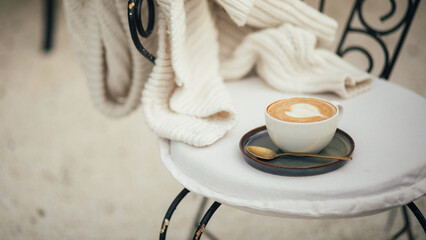 Cup of hot cappuccino on a chair near white knitted sweater.