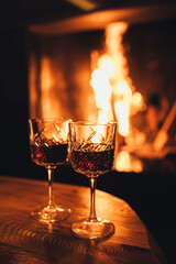 Two glasses of red wine on wooden table with cozy fireplace flame on the background.