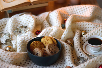 Cups of tea or coffee, bowl of cookies, soft blanket, lit candles, books and various Christmas decorations on the table. Selective focus.
