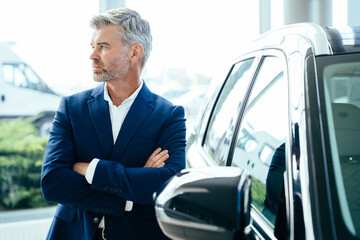 Serious mature grey hair beard businessman salesman wearing blue formal suit standing next to new car in showroom. - Powered by Adobe