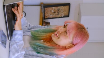 Happy diverse female with rainbow colored hair typing text on computer and laughing in a vertical 4K video clip. Portrait of a cheerful young woman from Middle Asia working on a laptop at home - Powered by Adobe