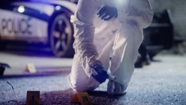 Forensics Technician in Coveralls Inspecting Potential Incriminating Evidence on Crime Scene. He Picks up a Marked Bullet Shell Clue and put it in Plastic Bag for Reservation. Police Gathering Proofs