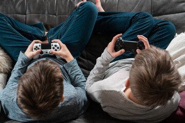 Guys sitting on sofa with game joysticks, from above view of crown and occiput. Children play videogames at home.