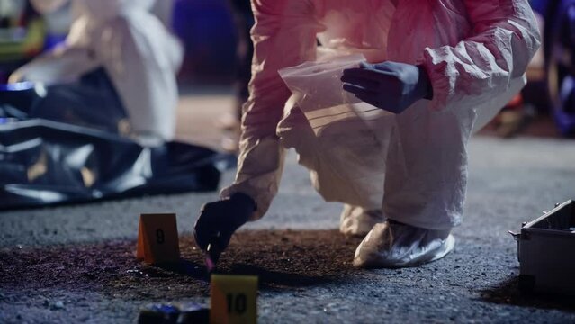 Forensics Unit Specialist Finding The Missing Murder Weapon on a Crime Scene. Man in Coverall Suit Observing Blood Patterns on the Knife After Packing it in a Sterilized Plastic Bag as Evidence.