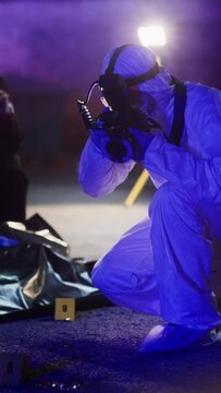 Vertical Shot: Forensics Specialist Taking Photos of Marked Evidence on a Crime Scene at Night. Expert Finds Glasses Potentially Belonging to the Dead Victim and Professionally Takes Photos of it
