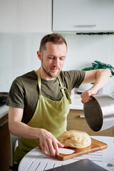 Making pastry at home kitchen concept. Bearded male chef baker in green apron getting out dough from metal bowl on a wooden cutting board using plastic spatula. High quality image