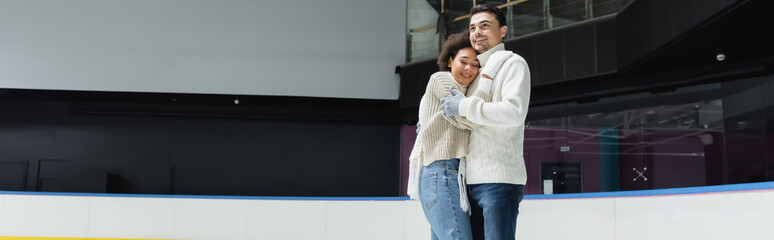 Man in knitted sweater hugging african american girlfriend on ice rink, banner