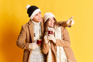 Portrait of stylish young man and woman in warm winter clothes posing, drinking mulled wine isolated over yellow background