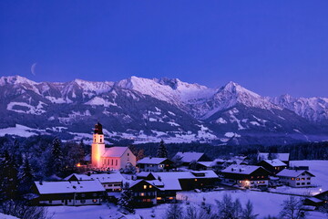 Bergdorf in den Alpen im Winter mit Abendstimmung in den Bergen und Mondsichel