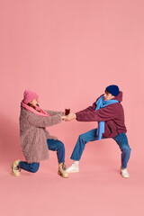 Portrait of young man and woman in knitted hat and scarf posing, fighting for mulled wine glass isolated over pink background