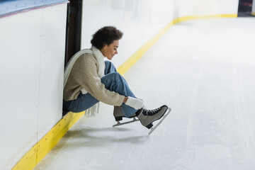 Side view of smiling african american woman wearing ice skates on rink - obrazy, fototapety, plakaty