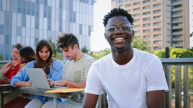 On foreground a smiling cheerful young African american student with a positive smile, white perfect teeth, looking at camera sitting outside taking a break at the university. at the background a