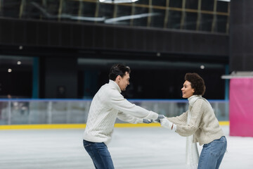 Side view of young interracial couple holding hands while ice skating on rink