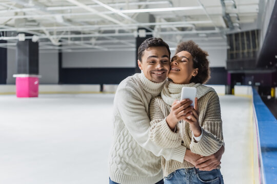 Positive man in sweater hugging african american girlfriend with smartphone on ice rink