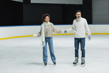 Smiling multiethnic couple in sweaters and jeans skating during date on ice rink