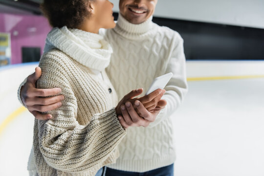 Cropped view of young multiethnic couple in warm clothes hugging and using smartphone on ice rink