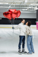 Side view of smiling man holding balloons in shape of heart near african american girlfriend on ice rink