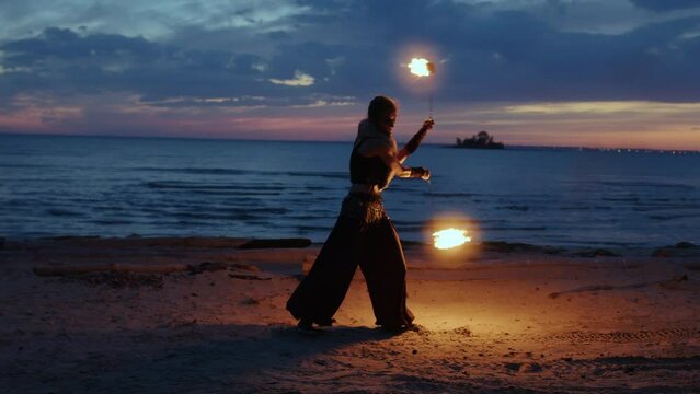 Girl in tribal fusion costume turns around herself, rotating burned sticks in hands on river bank. Fire dancer twirling with torches. Fire performance on night seashore. Young woman moving in fireshow