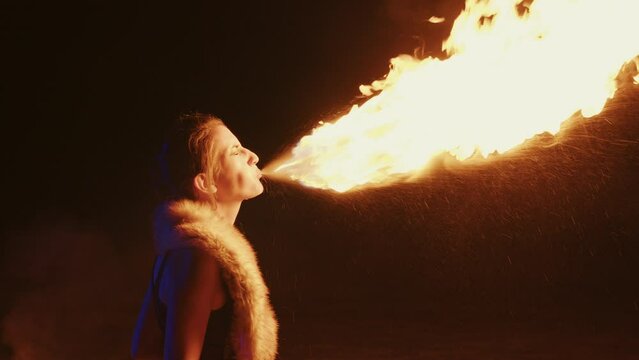 Young woman performing fire breathing using burning torch at night. Fire show artist lets out fiery cloud from mouth in open-air. Fire breather girl ignites flame at evening street show. Profile view