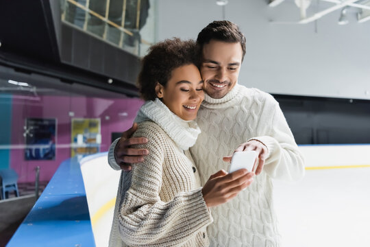 Smiling man hugging african american girlfriend and using smartphone on ice rink