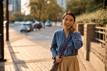 Happy Asian businesswoman talking on cell phone while walking on street.