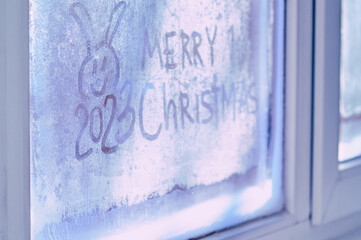 Merry Christmas on a frozen window. Rabbit symbol of Chinese New Year 2023. Merry Christmas and Happy New Year greeting concept. Sunlight through frosty patterns. Selective focus