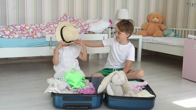 Happy children in white clothes and straw hat pack the luggage suitcase for travel indoor children room. Concept of family beach vacation, preparation, international trip journey.