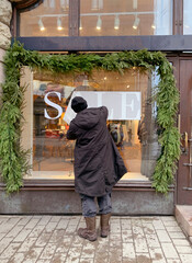 worker sticks word sale, winter street Helsinki city, green garland decorates shop window, concept...