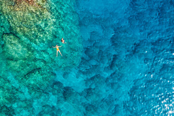 Aerial view of a young couple snorkeling above coral reef reaching deeper parts of the crystal clear water, Rhodes, Greece. Aerial drone view of a couple snorkeling swimming in crystal clear water. - obrazy, fototapety, plakaty
