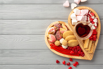 Valentine's Day concept. Top view photo of wooden heart shaped serving tray with confectionery chocolate jelly candies cookies and glass cup of drinking on grey wooden desk background with empty space