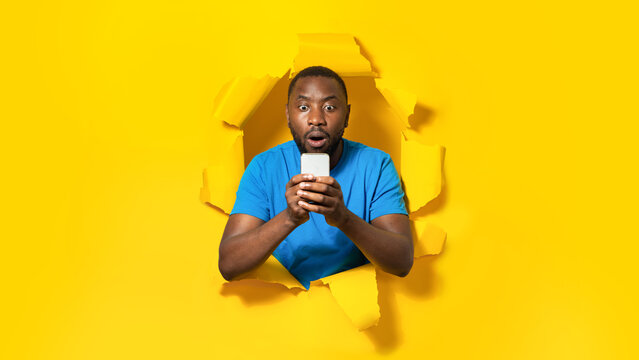 Shocked Black Man Using Cellphone, Reading Shocking Message, Posing In Hole Of Torn Paper On Yellow Background