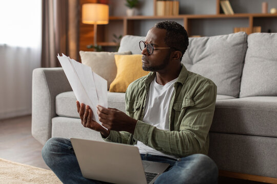 Sad pensive mature black man in glasses with laptop analyst document, work in living room interior