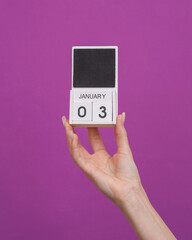 Female hand holding wooden calendar with date January 03 on purple background
