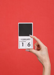 Female hand holding wooden calendar with date February 16 on red background